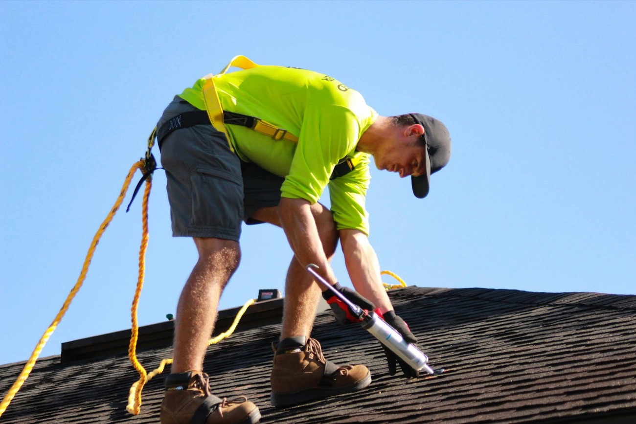A roofer in a safety harness nailing shingles on a house roof